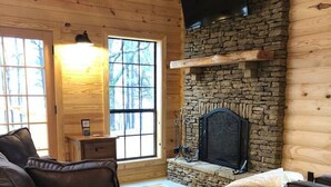 A log cabin interior with a stone fireplace, leather sofas, and a large window.