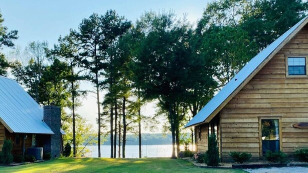 A cabin with a metal roof, a wooden deck, and a view of a lake surrounded by trees.