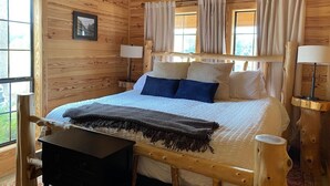 A wooden cabin bedroom with a bed, two windows with curtains, a desk, and a chair.