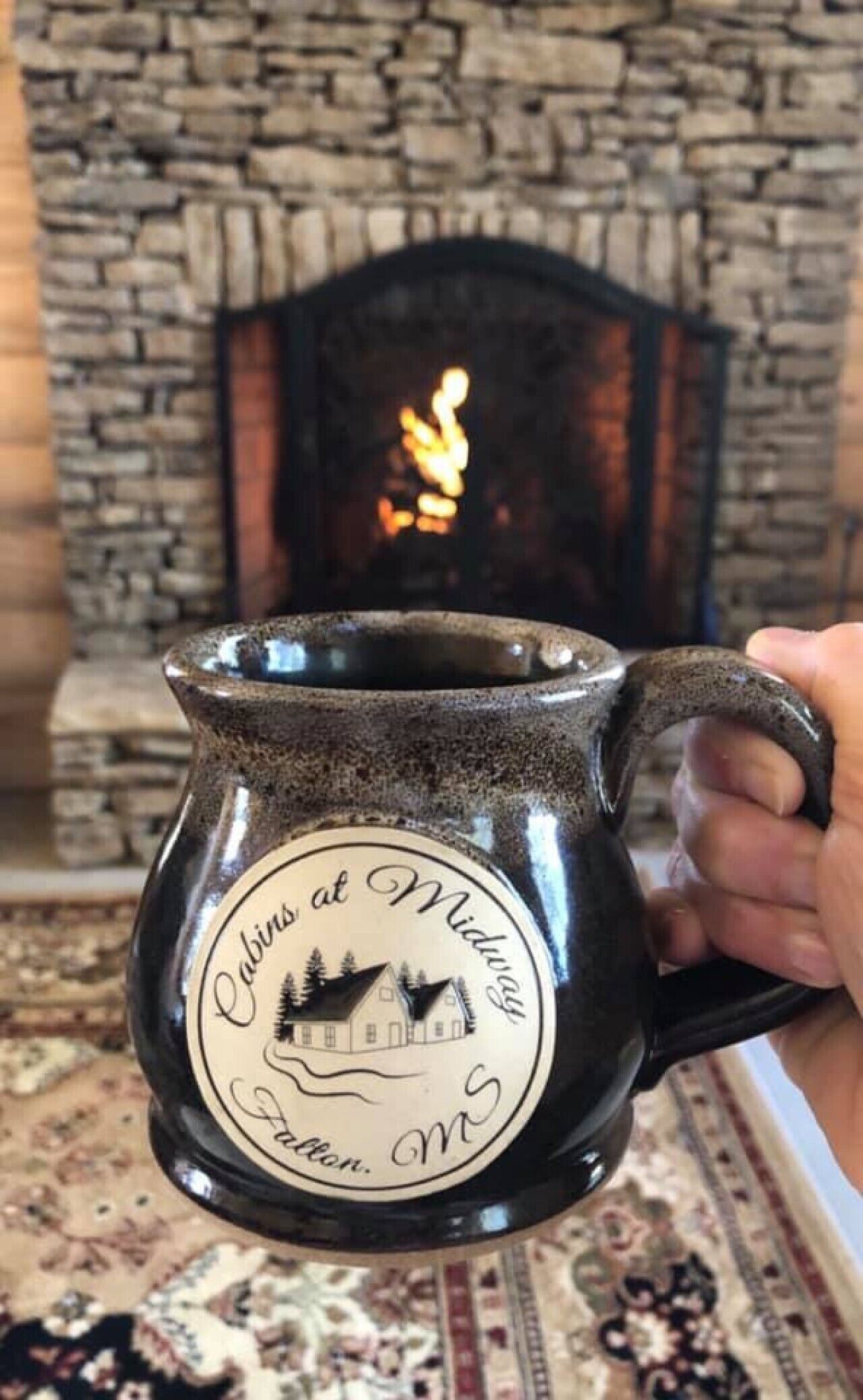A hand holding a ceramic mug with a label that reads 'Cabins at Midway, MS.'