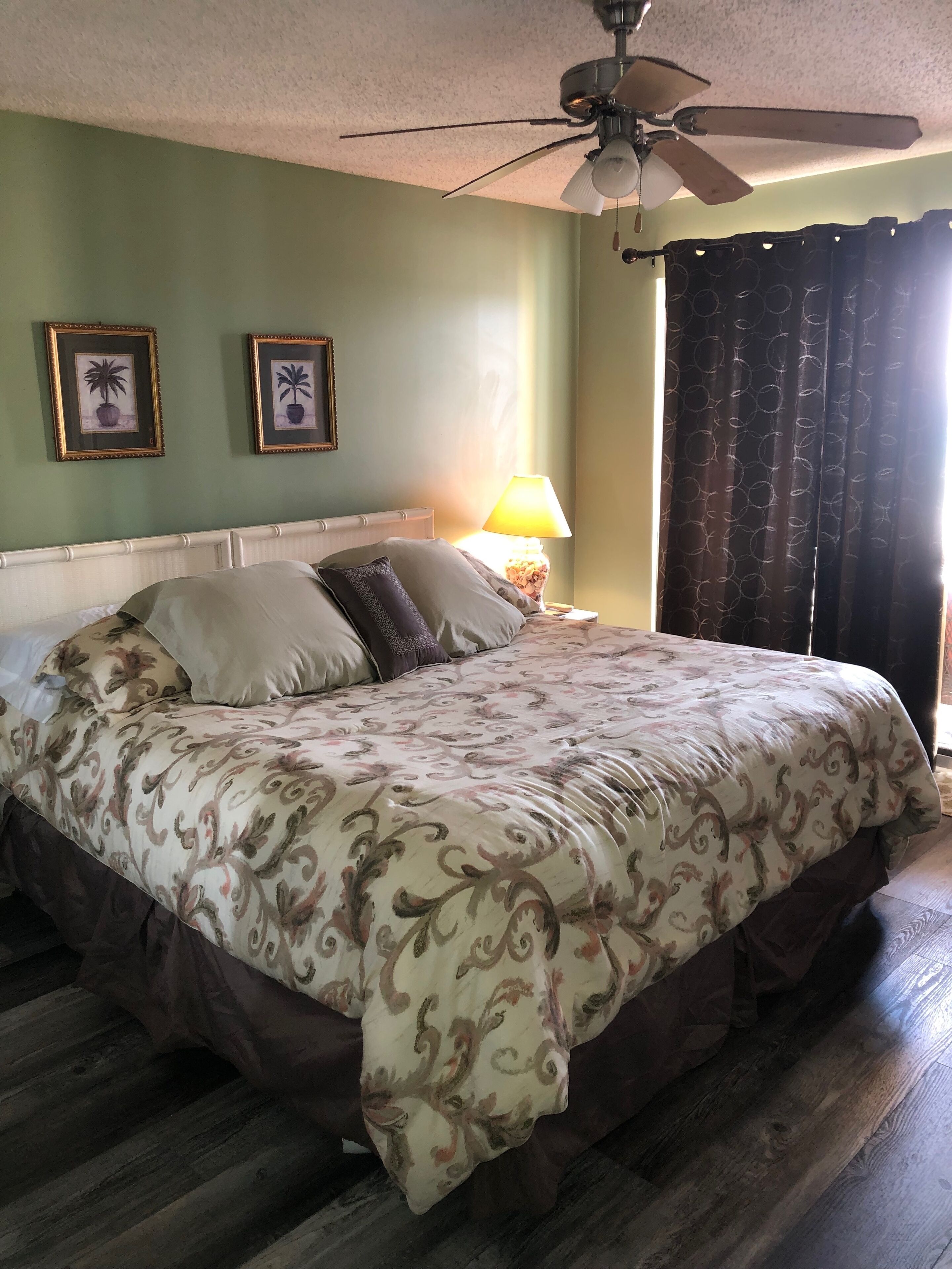 Un dormitorio con una cama, un ventilador de techo, una mesita de noche con lámpara y dos cuadros en la pared.