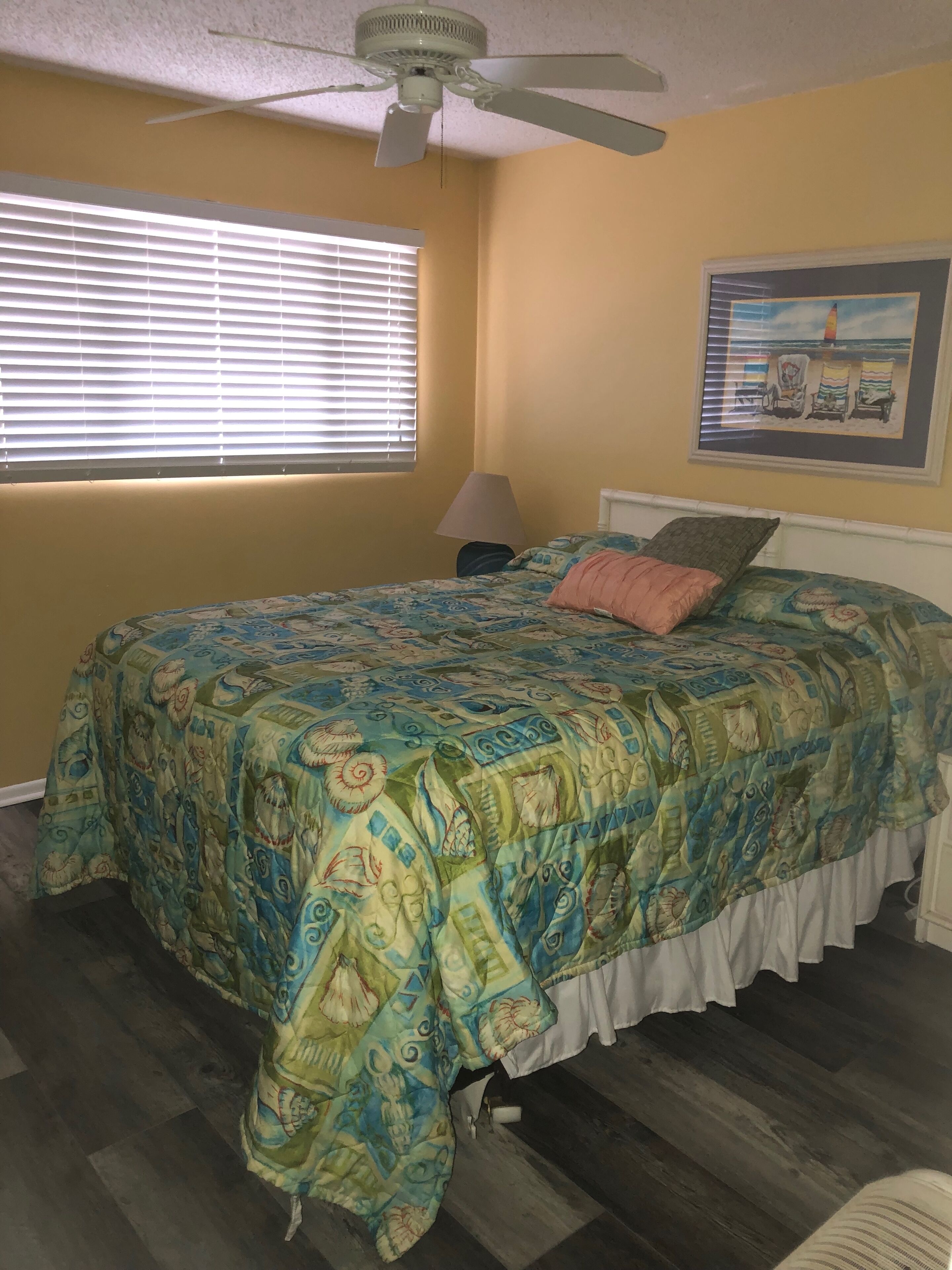 A bedroom with a bed, a ceiling fan, a window with blinds, a lamp, and a beach-themed painting on the wall.