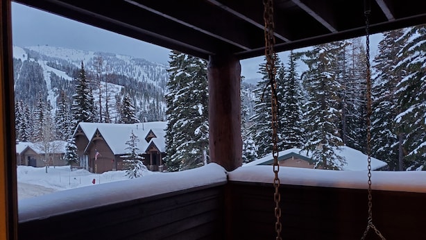 A wooden cabin with a porch, a swing, and a view of snow-covered mountains and trees.