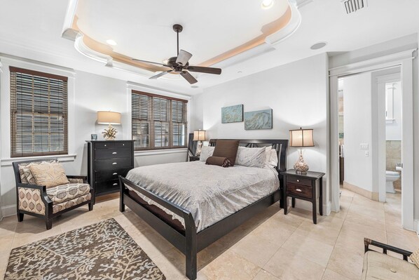 A bedroom with a bed, a chair, a nightstand, a ceiling fan, and two windows with blinds.