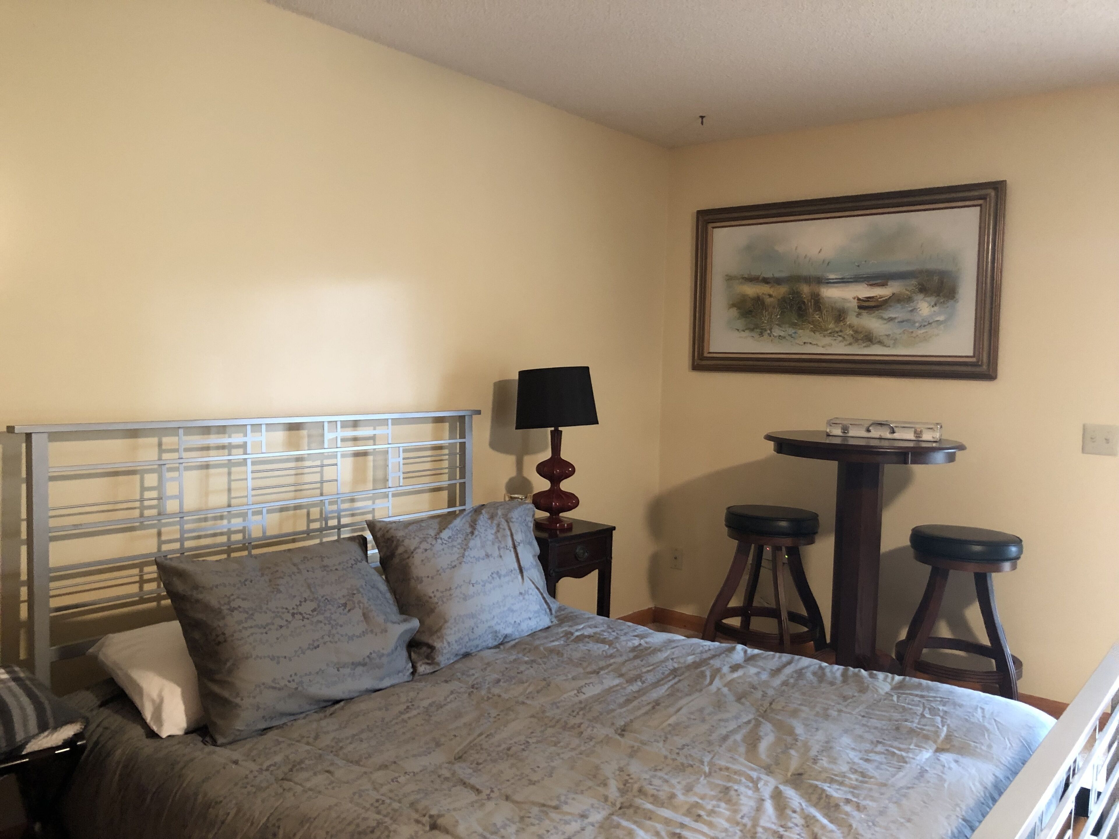 A bedroom with a bed, a nightstand, a lamp, a small table, and a stool.