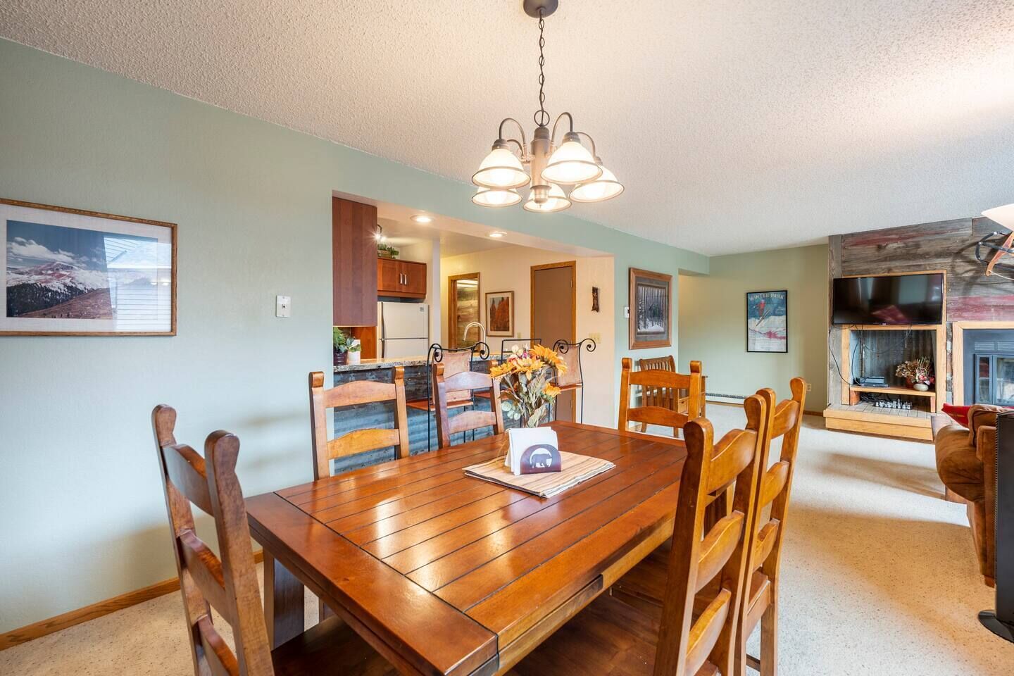 A dining area with a wooden table and chairs, a chandelier, and a kitchen in the background.