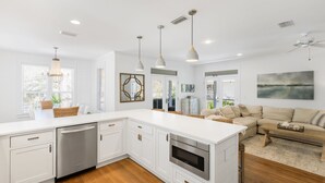 A modern kitchen with white cabinetry, a built-in oven, and a large island. There is a dining area with a table and chairs, and a living room with a sofa and a coffee table. - Hakuna Matata - 2 Community Pools, Tennis, & Bikes (Santa Rosa Beach)