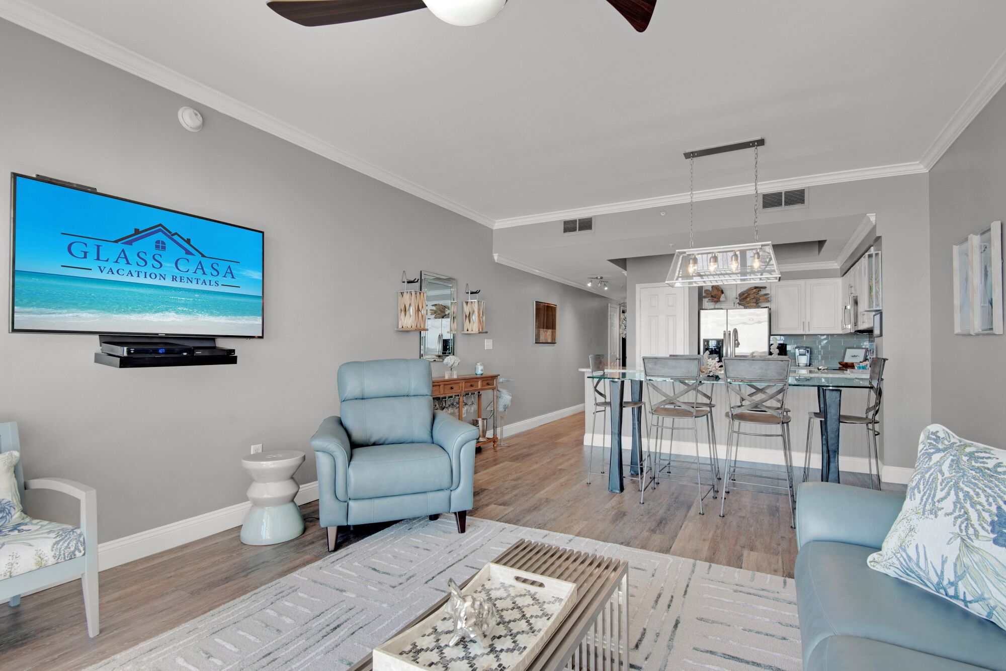 Una sala moderna con televisor, un sillón azul, una mesa de centro de vidrio y un comedor con mesa y sillas de vidrio.