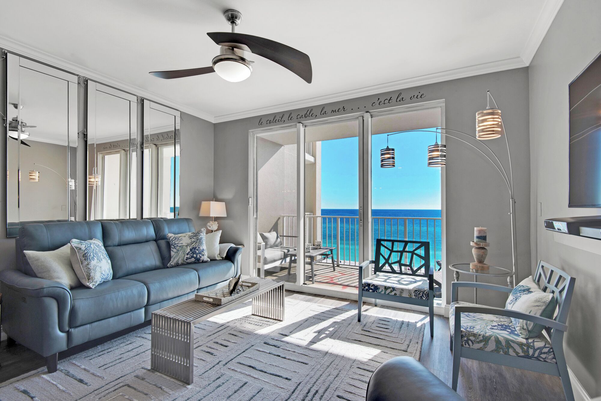 A modern living room with a grey sofa, a coffee table, and a balcony with a view of the sea.