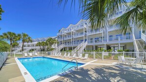 Una piscina con valla blanca, un edificio de varios pisos con balcones y palmeras.