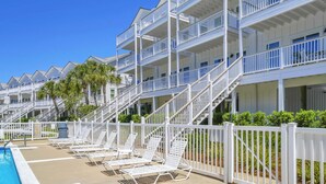 Un hotel de varios pisos con balcones, una zona de piscina con tumbonas y un cielo azul despejado.