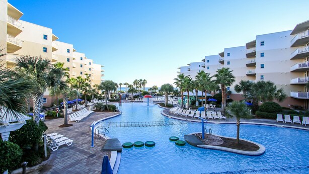 Zona de piscina en un resort, con sillas de descanso, palmeras y cielo azul brillante.