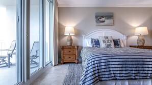 Un dormitorio con un amplio ventanal corredizo, una cama con ropa de cama a rayas, dos mesitas de noche con lámparas y una almohada con motivos playeros.