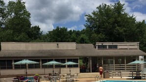 Una piscina con sillones de descanso y sombrillas, un área de estar techada y un edificio con techo plano y ventanas.