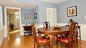A dining room with a wooden table and chairs, a window with blinds, and framed pictures on the wall.