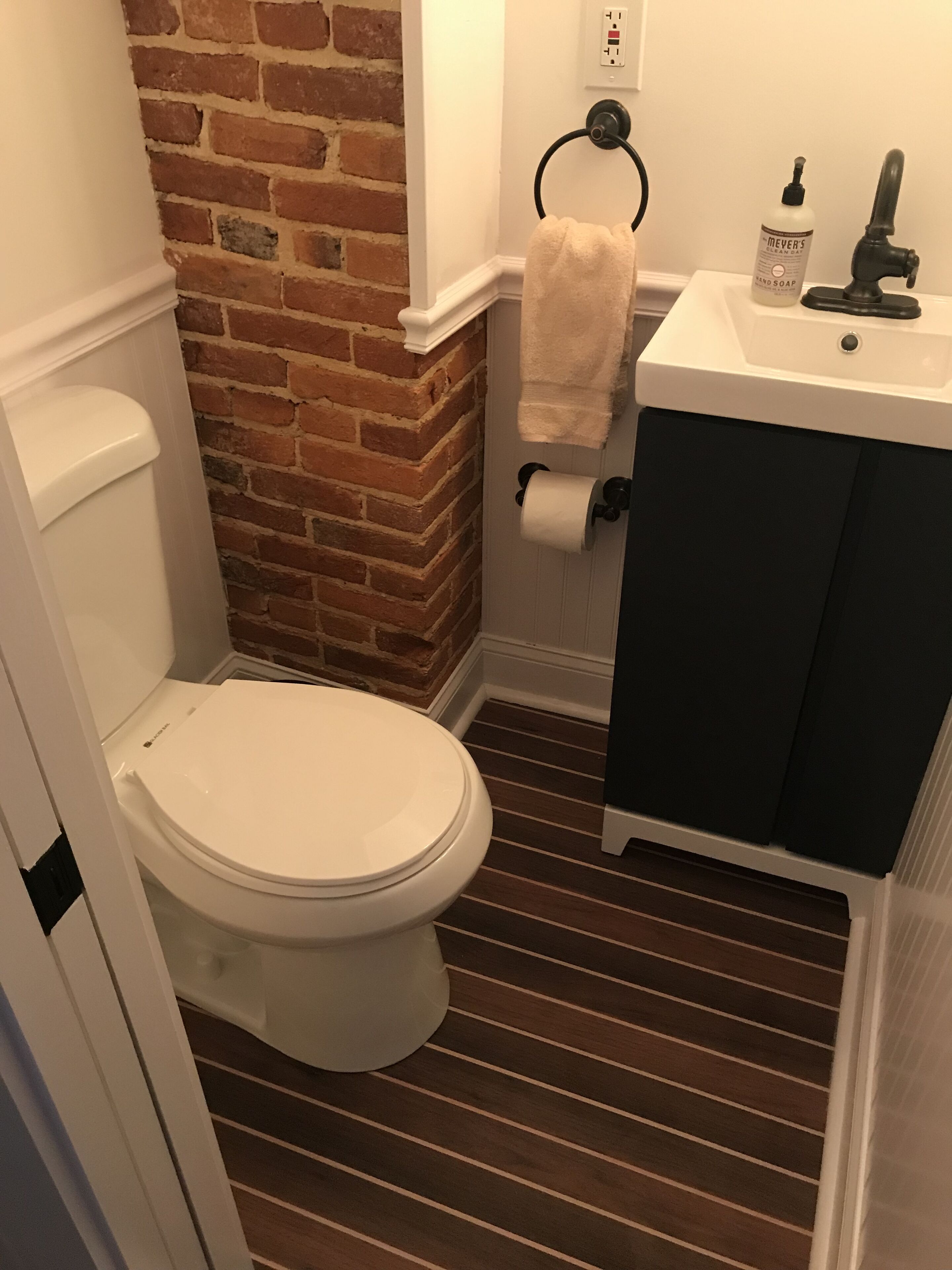 Une salle de bain avec des toilettes blanches, un mur d’accent en briques, un sol à rayures, un lavabo blanc avec un meuble noir et un distributeur de savon pour les mains.