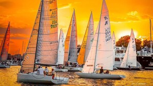 A fleet of sailboats with registration numbers and flags, docked at a marina during sunset.