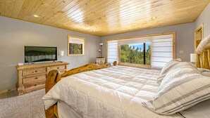 A bedroom with a large bed, wooden headboard, a dresser, a television, and a window with a view of trees.