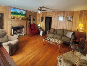 A living room with wooden paneling, a fireplace, a red armchair, a green sofa, and a wooden coffee table. - The Farm House at Peaks of Otter Winery and Orchards (Bedford)
