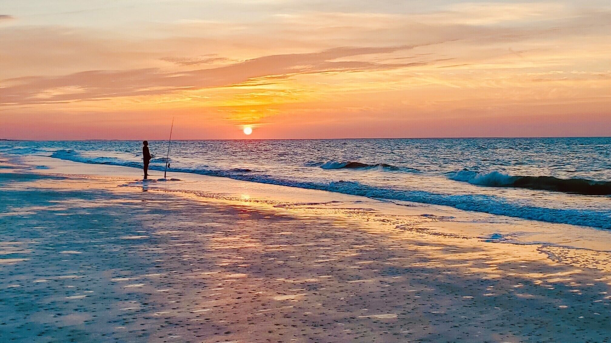 A beach with a sunset, waves, and a person fishing.