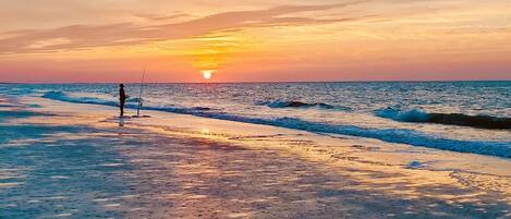 A beach with a sunset, waves, and a person fishing.