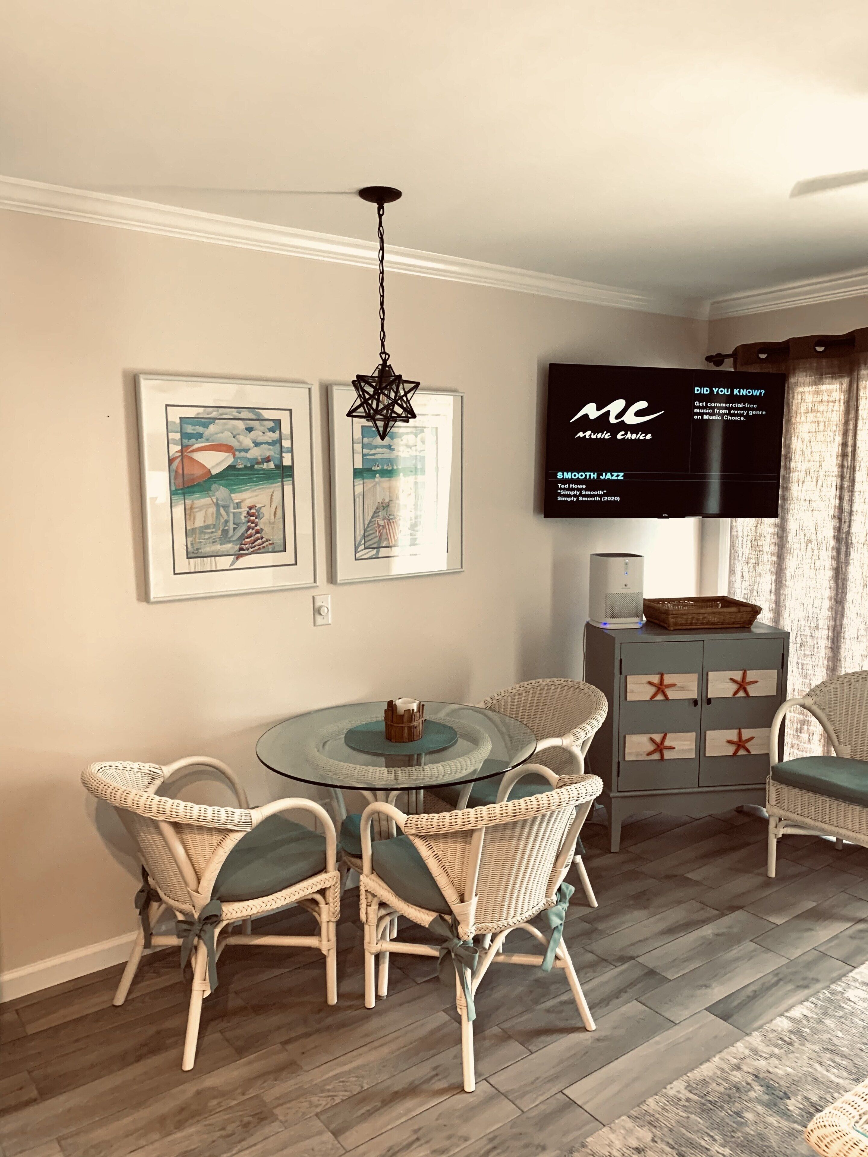 A dining area with a round glass table, wicker chairs, a starfish pendant light, and a flat-screen TV mounted on the wall.