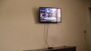 A television mounted on a wall above a dark wooden dresser with a trash can beside it.