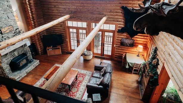 A log cabin interior with a stone fireplace, wooden floors, a dining area, and a mounted deer head.