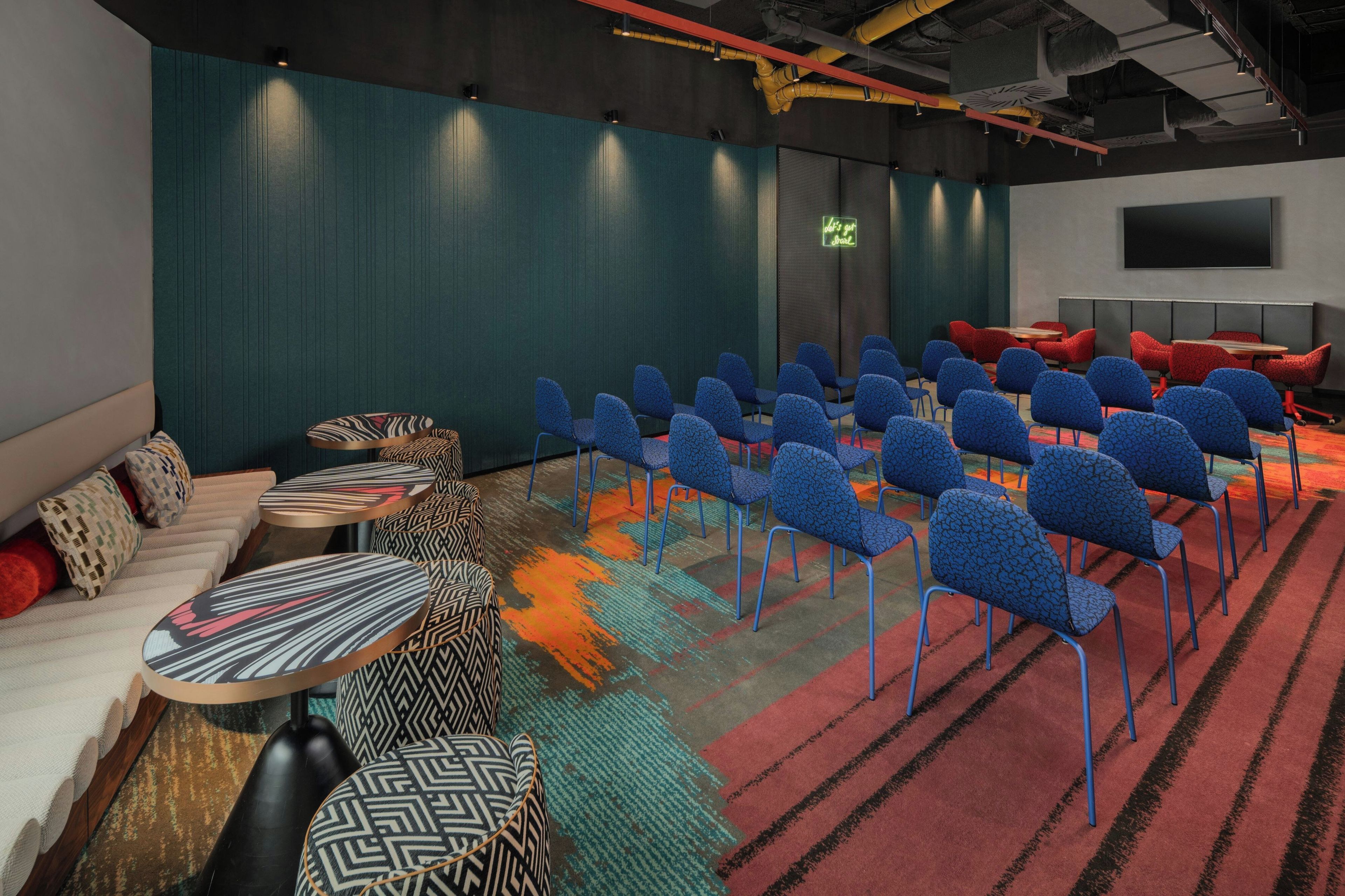 A conference room with rows of blue chairs, a sofa, and patterned ottomans.