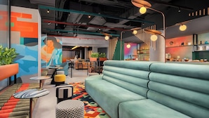 Een moderne zithoek met een lichtblauwe bank, een kleurrijk geometrisch vloerkleed en diverse zitplaatsen, waaronder een gestreept poef en een ribbelbank.