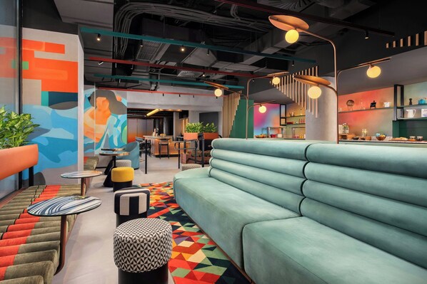 A modern lounge area with a light blue sofa, colorful geometric rug, and a variety of seating options including a patterned ottoman and a striped bench. - Aloft by Marriott Madrid Gran Via (Madrid)