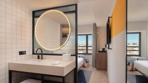 Een moderne hotelbadkamer met een grote spiegel, een wastafel en een badkamermeubel. Op de achtergrond is een bed met uitzicht op de stad te zien.
