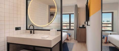 Un moderno baño de hotel con un espejo grande, un lavabo y tocador. Se ve una cama con vista a la ciudad al fondo.