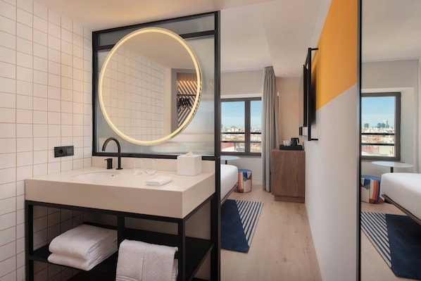 A modern hotel bathroom with a large mirror, a sink, and a vanity. There is a bed with a view of the city in the background.