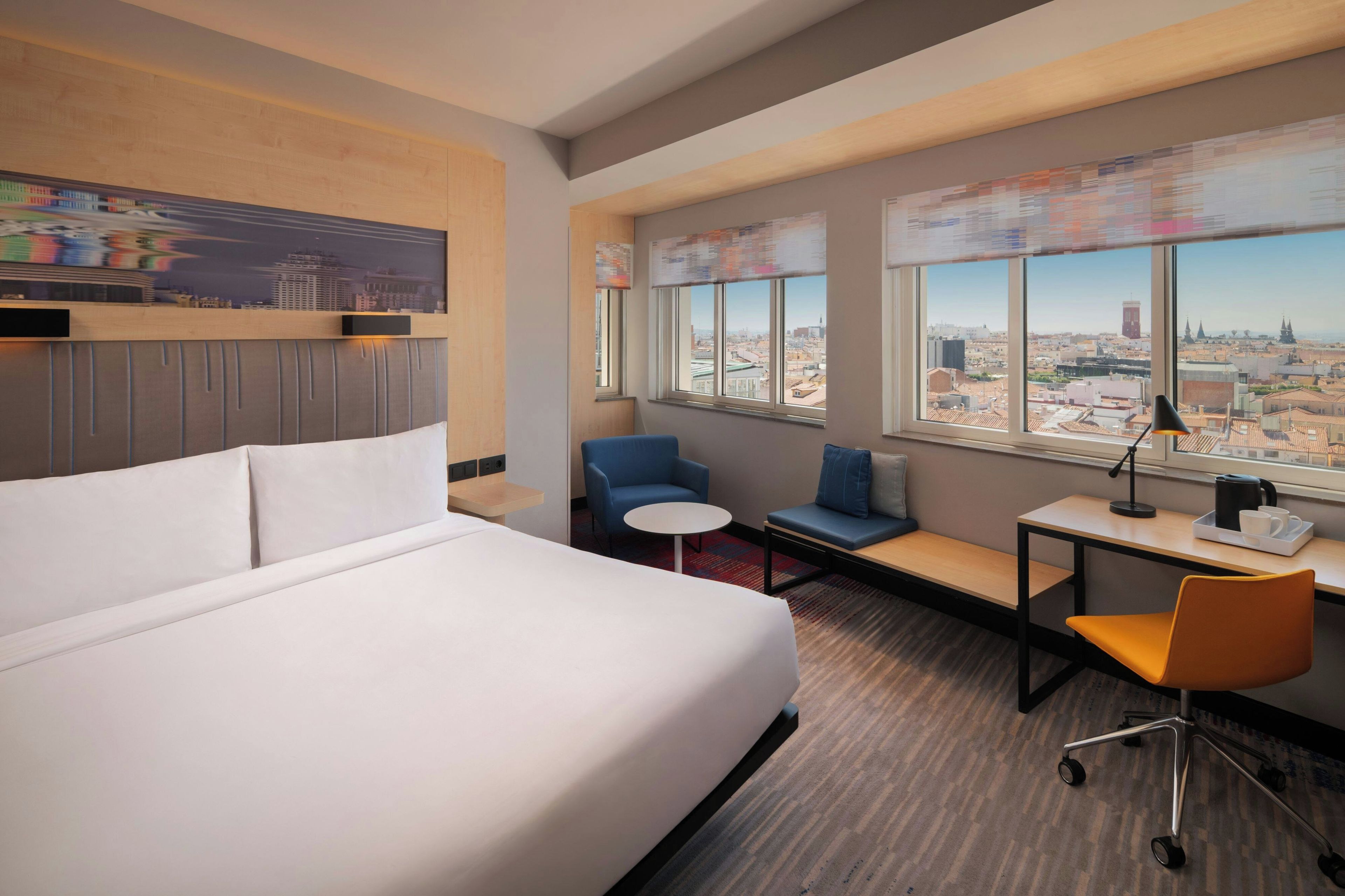 Ein Hotelzimmer mit einem großen Bett, einem Schreibtisch, einem Stuhl und einer Couch. Große Fenster bieten Blick auf die Stadt.