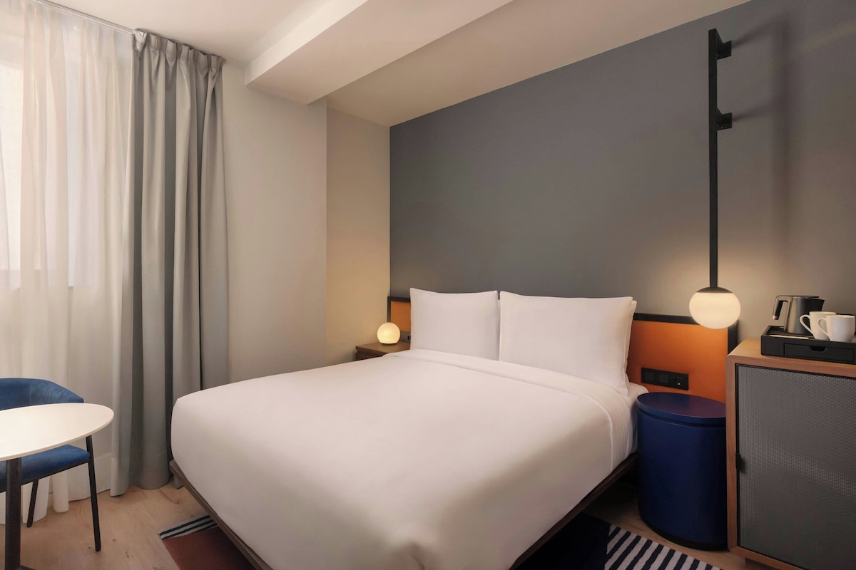 Una habitación de hotel con una cama, un escritorio, una silla, una mesita de noche con lámpara y un armario pequeño.