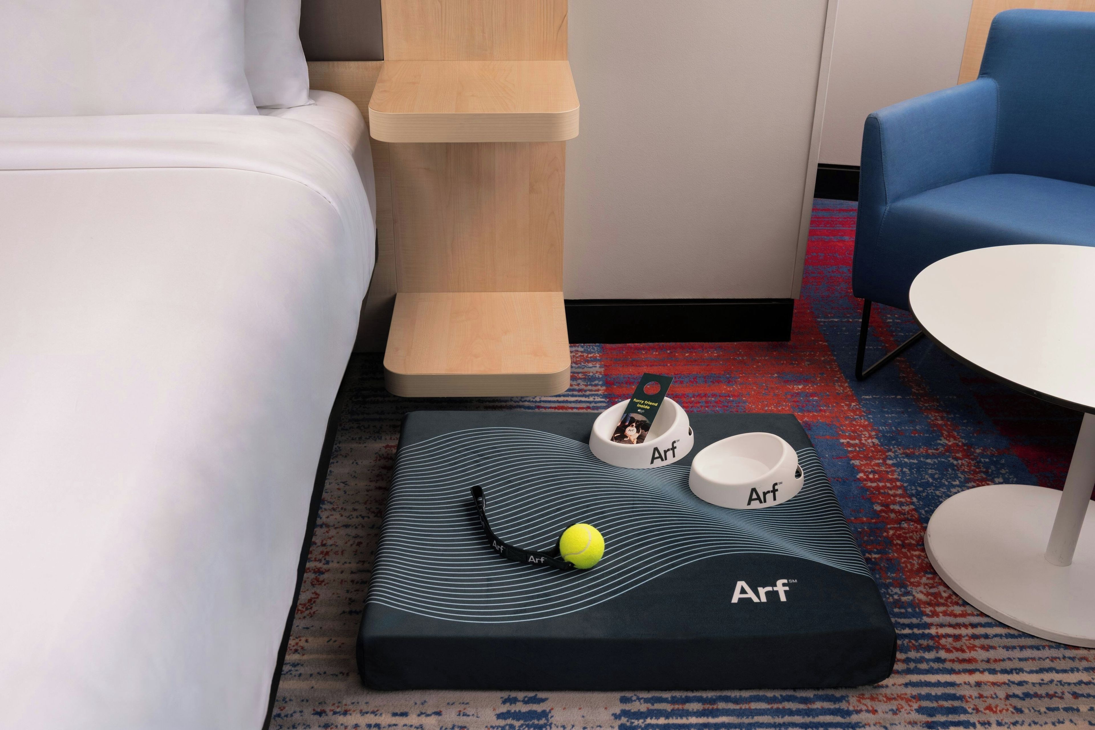 A hotel room with a bed, a small table with a tennis ball and a card, a blue chair, and a white table.