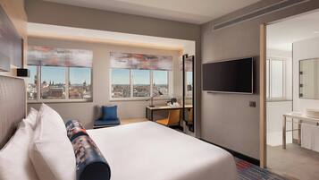 Ein Hotelzimmer mit einem großen Bett, einem Schreibtisch, einem Sessel, einem Fernseher und Blick auf die Stadt.