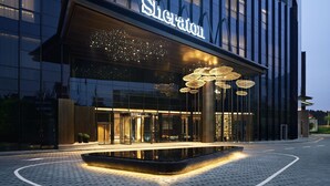 Una entrada de hotel moderna con fachada de vidrio, señalización iluminada y una fuente de agua al frente.