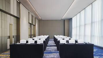 Un salón de conferencias con filas de sillas y mesas blancas, ventanas amplias y suelo alfombrado.