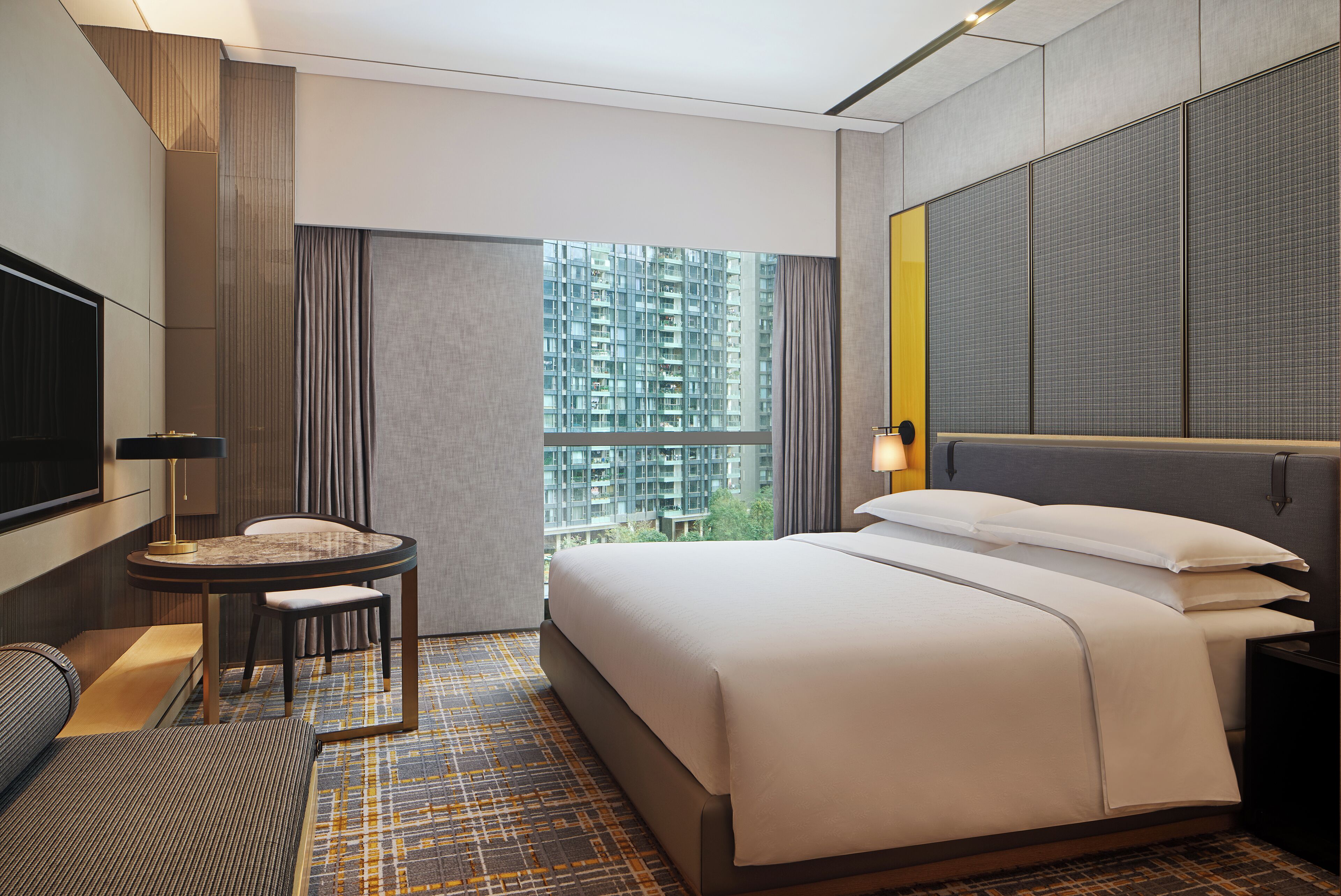 A modern hotel room with a large bed, a desk, and a view of a high-rise building.