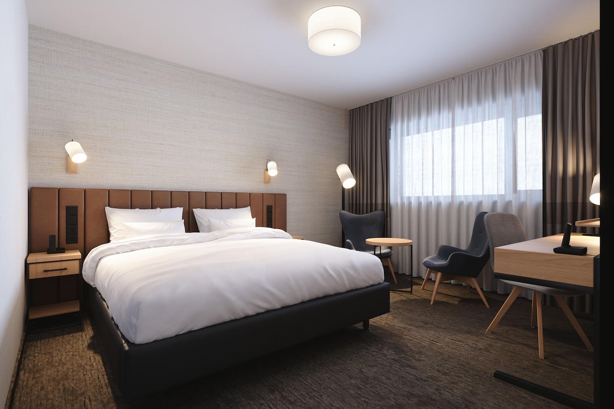 A modern hotel room with a large bed, a desk, two chairs, and a small table.