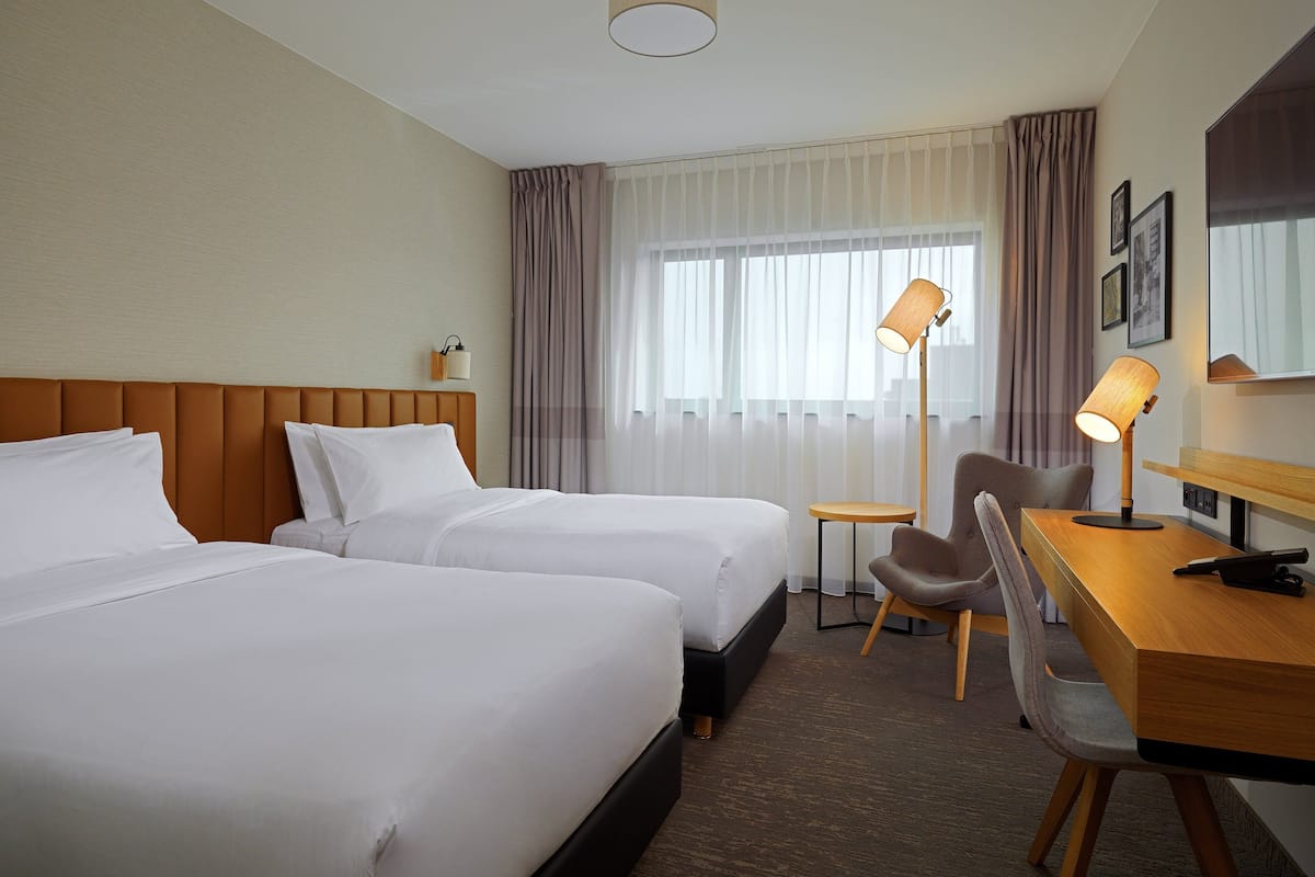 A hotel room with two beds, a desk with a chair, a lamp, and a large window with curtains.