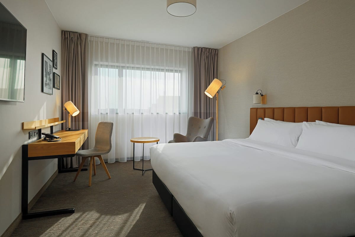 A hotel room with a large bed, a desk with a chair, a small table, and a lamp.