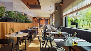 A restaurant with a long counter, multiple tables, and chairs. There are potted plants on the left side and a window with a view of trees. - Four Points by Sheraton Warsaw Mokotow (Warsaw)