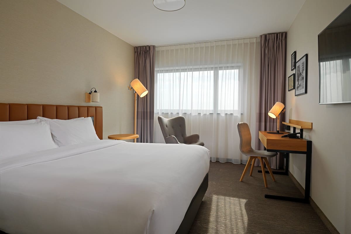 A hotel room with a large bed, a desk, two chairs, and a window with curtains.