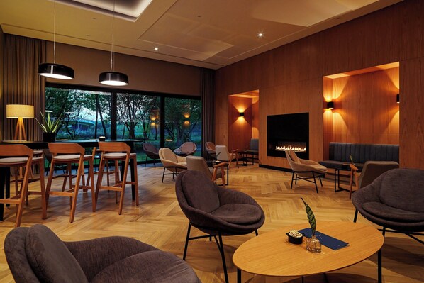 A modern hotel lobby with a fireplace, comfortable seating, and large windows offering a view of trees.