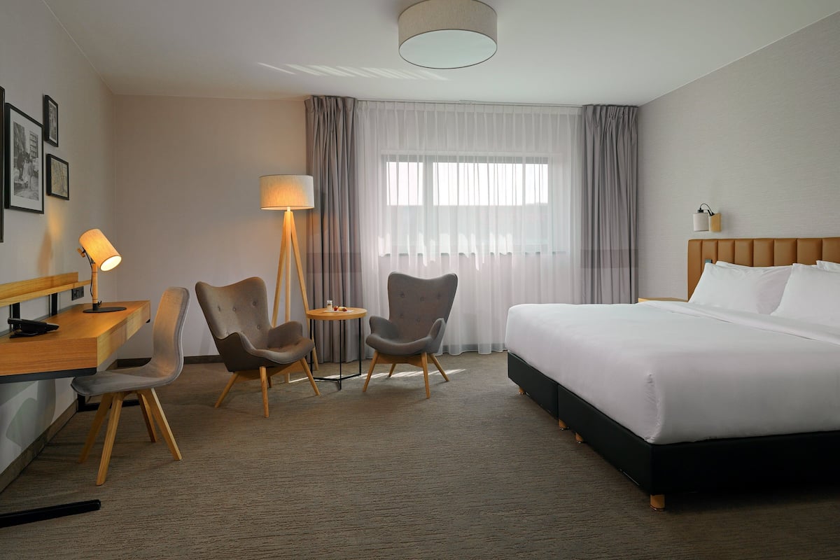 A hotel room with a bed, two armchairs, a desk, a lamp, and a window with curtains.