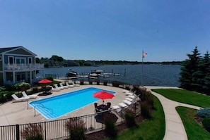 A swimming pool with lounge chairs and umbrellas, a dock with boats, and a view of a body of water.