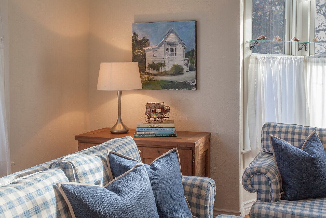 Un salon chaleureux avec un canapé à carreaux, une lampe, un tableau représentant une maison et une fenêtre avec des rideaux.